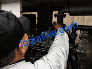 Technician in a white coverall adjusting a large industrial engine inside a machinery compartment, wearing protective glasses and a cap, viewed from behind.