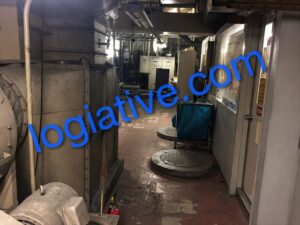 地下搬出通路_R / 非常用発電機の専門会社 株式会社ロジエイティブ Industrial machine room corridor with large metal cabinets, wet floor, and a blue cart against the right wall in the background.